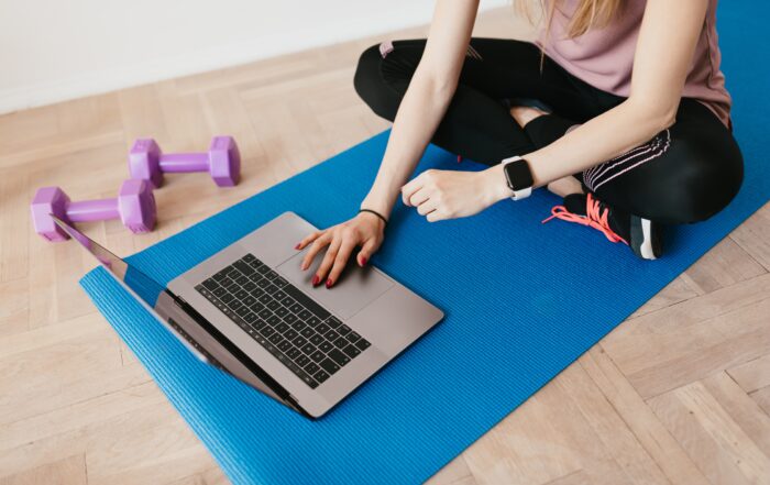Auf einer Yoga-Matte sietzt eine weiblich gelesene Frau vor einem Laptop, der auch auf der Matte liegt. Daneben liegen zwei Gymnastikhanteln. Es wird die Assoziation Fittness und digitale Medien angesprochen.