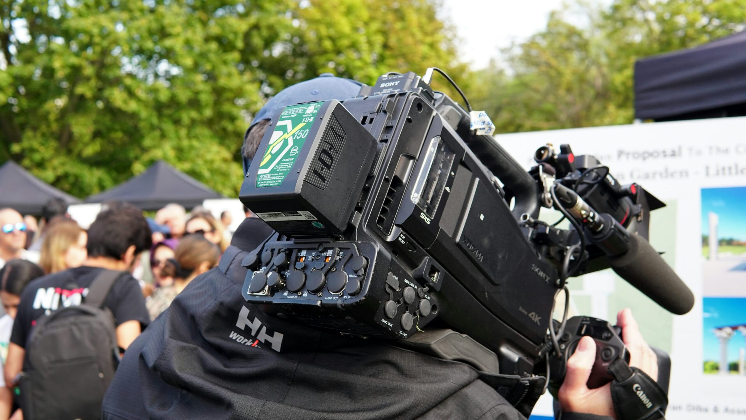 Eine Person filmt mit einer professionellen Videokamera eine Veranstaltung im Freien; im Hintergrund stehen mehrere Menschen.